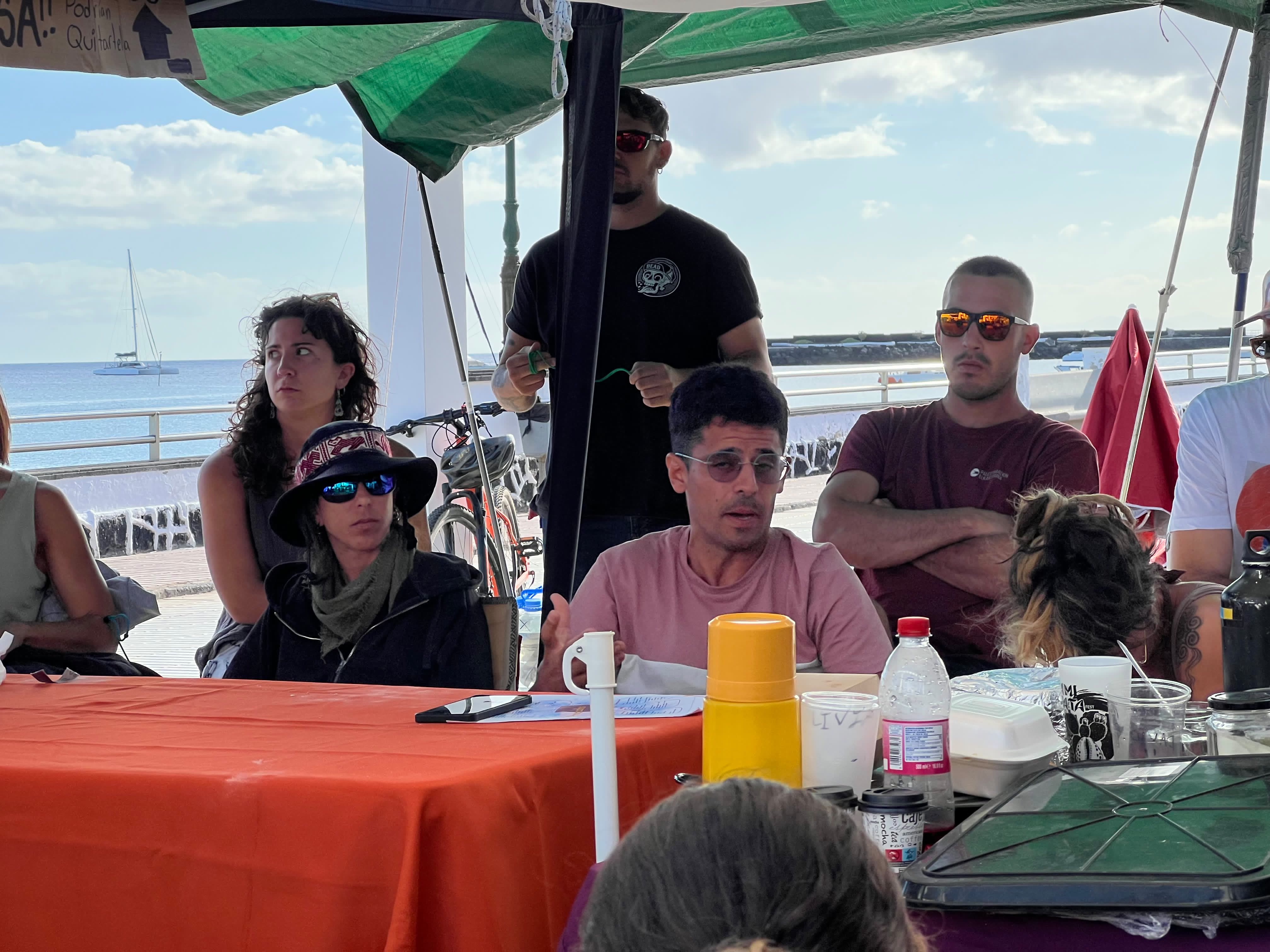 Personas en la mesa de la asamblea bajo la carpa, con el puerto detrás